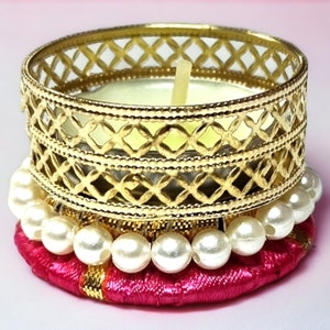 May include: A decorative candle holder with a gold filigree design. The candle holder sits atop a pink, embroidered base adorned with white pearls. A lit candle is inside the holder.