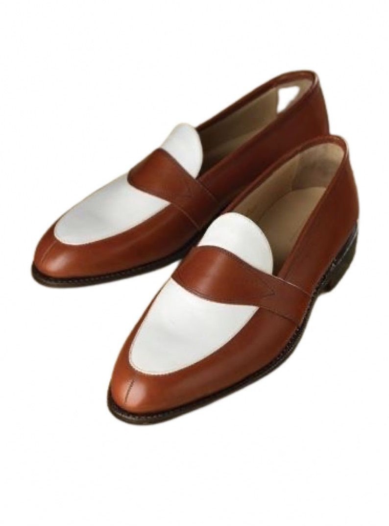 May include: A pair of brown leather loafers with white leather accents. The loafers have a rounded toe and a slip-on design.