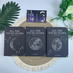 May include: Three hardcover books titled "ALL THE YOUNG DUDES" with celestial illustrations on a dark background. The books are arranged with a matching bookmark. The book spines have gold accents. The books are on a beige circular mat.