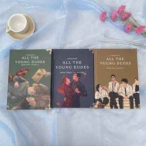 May include: Three hardcover books titled "ALL THE YOUNG DUDES" are displayed. The covers feature illustrations and text, with gold corner accents. The books are in shades of green, navy, and brown. A teacup and saucer are in the upper left corner, and pink flowers are in the upper right.