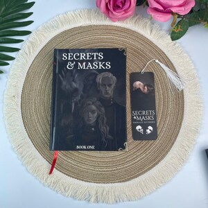 Peut inclure: Un livre intitul&eacute; "Secrets & Masks: Book One" avec un marque-page. La couverture du livre pr&eacute;sente une illustration sombre de deux personnes. Le marque-page porte le m&ecirc;me titre et une gland. Les articles sont sur un dessous de plat beige.