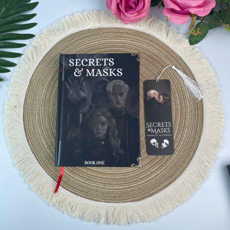 Peut inclure: Un livre intitul&eacute; "Secrets & Masks: Book One" avec un marque-page. La couverture du livre pr&eacute;sente une illustration sombre de deux personnes. Le marque-page porte le m&ecirc;me titre et une gland. Les articles sont sur un dessous de plat beige.