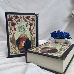 May include: Two hardcover books with black covers and decorative art. The covers feature a couple kissing, surrounded by floral designs and the text "Amor Vincit Omnia". The books have gold corner protectors.