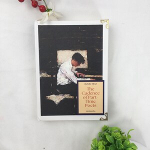 May include: A book titled "The Cadence of Part-Time Poets" with cover art depicting a person at a desk. The cover has a dark background and a figure in white. Gold corner protectors are visible.
