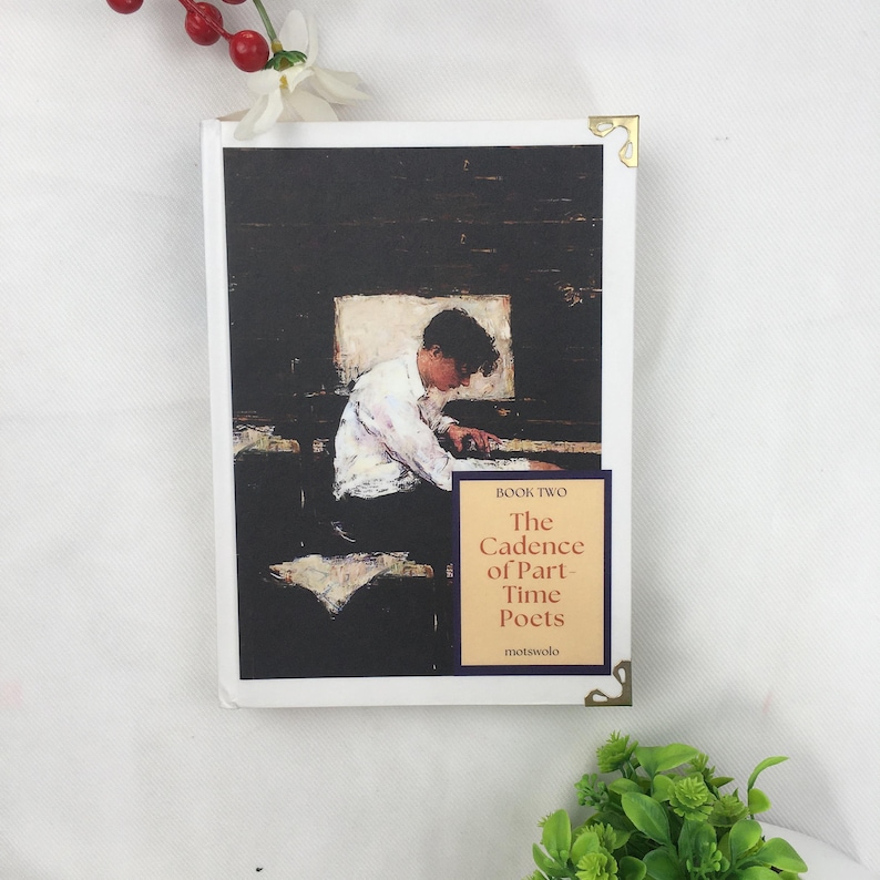May include: A book titled "The Cadence of Part-Time Poets" with cover art depicting a person at a desk. The cover has a dark background and a figure in white. Gold corner protectors are visible.
