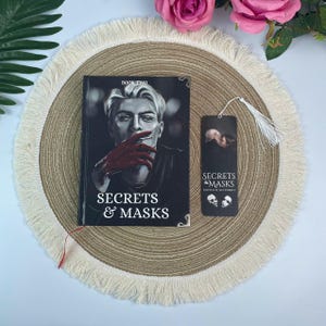 Peut inclure: Un livre intitul&eacute; "Secrets & Masks" avec une couverture repr&eacute;sentant un homme avec des gants rouges. Un marque-page assorti avec un pompon est &agrave; c&ocirc;t&eacute; du livre. Le livre et le marque-page sont sur un dessous de plat rond et &agrave; franges.