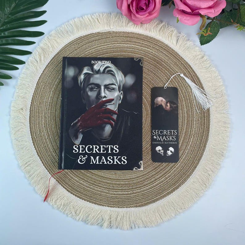 Peut inclure: Un livre intitul&eacute; "Secrets & Masks" avec une couverture repr&eacute;sentant un homme avec des gants rouges. Un marque-page assorti avec un pompon est &agrave; c&ocirc;t&eacute; du livre. Le livre et le marque-page sont sur un dessous de plat rond et &agrave; franges.