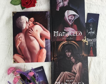 May include: A collection of fantasy novels titled "Manacled" with various cover art featuring illustrations of people. The covers are black with gold corner accents. A bookmark with the same title is also present. A red rose and pink flowers are in the background.
