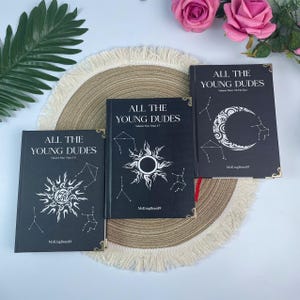May include: Three black books with gold corner detailing are displayed on a round, fringed mat. The books, titled "ALL THE YOUNG DUDES," feature white celestial designs and text. The books are labelled Volume One, Two, and Three.