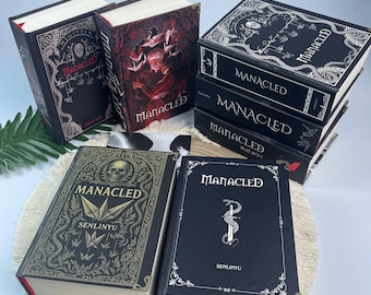 May include: A collection of hardcover books with the title "Manacled" in a gothic font. The books have black covers with ornate gold and white designs, including skulls, snakes, and other illustrations. Some books are stacked, others are lying flat.