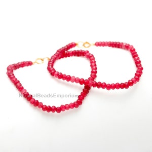 May include: Two red beaded bracelets with gold-colored clasps are displayed against a white background. The beads are small and faceted, creating a sparkling effect. The bracelets are connected by a gold-colored clasp.