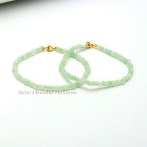 Op de afbeelding: Twee delicate armbanden met kleine, facetgeslepen, lichtgroene kralen. Elke armband heeft een goudkleurige sluiting. De armbanden worden gepresenteerd op een witte ondergrond. De tekst "NaturalBeadsEmporium" is zichtbaar onderaan.