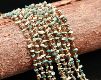 Può includere: Primo piano di una collana di perline a più fili. La collana presenta piccole pietre turchesi e bianche di forma irregolare, intervallate da perline dorate. I fili sono drappeggiati su un pezzo di legno marrone strutturato.