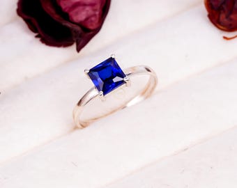 May include: A silver ring with a square, deep blue gemstone. The ring is set on a white surface, with dried burgundy flower petals in the background. The gemstone is the focal point, showcasing its vibrant color and cut.
