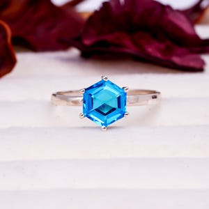 May include: A silver ring with a large, hexagonal, bright blue gemstone. The gemstone is faceted, catching the light and creating a sparkling effect. The ring band is thin and polished, complementing the vibrant blue of the stone. The ring is a piece of jewelry.