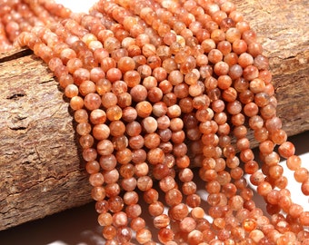 May include: Strands of sunstone beads in shades of orange and peach. The beads are round and have a speckled appearance. The beads are strung together, creating a flowing, textured effect. The beads are displayed on a piece of wood.