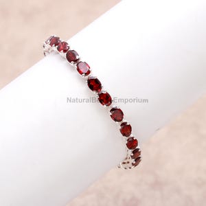 May include: A silver bracelet featuring a series of oval-cut, deep red gemstones. The gemstones are set closely together, creating a continuous band of color. The bracelet is displayed on a white surface.