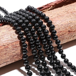 May include: Strands of glossy, black beads. The beads are round and uniform in size, with a polished finish. The beads are displayed on a piece of brown wood. The beads are strung together, suggesting they are for jewelry making.