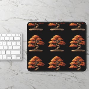 May include: Black rectangular mouse mat with a repeating pattern of bonsai trees. Each tree has a twisted trunk, orange leaves, and sits in a brown pot. The background is black. A white keyboard is visible on the left.