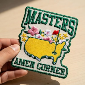 Puede incluir: Parche bordado con las palabras "MASTERS" y "AMEN CORNER" en letras verdes. El parche tiene un fondo beige con un mapa amarillo del campo de golf, una bandera roja y detalles florales en rosa, blanco y amarillo.