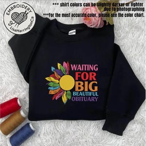 Op de afbeelding: Zwart sweatshirt met een kleurrijk zonnebloemontwerp en de tekst "WAITING FOR BIG BEAUTIFUL OBITUARY". De zonnebloem heeft gele, roze, oranje, blauwe en groene bloemblaadjes. Het sweatshirt heeft ook de tekst "EMBROIDERY LisaHarringtonCa".
