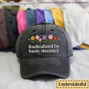 Op de afbeelding: Zwarte geborduurde baseballpet met de tekst "Radicalized by basic decency" en een rij kleurrijke bloemen. De pet is gemaakt van een distressed denim stof.