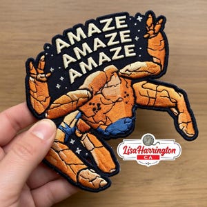 May include: Embroidered patch featuring a muscular figure with the word "AMAZE" repeated three times on a black background. The figure is orange and blue, with white text and star accents. The patch has a unique shape and a Lisa Harrington CA logo.