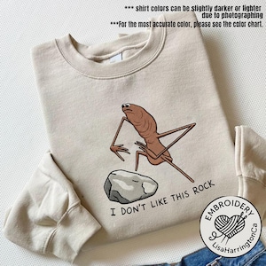 Puede incluir: Sudadera beige con un personaje de dibujos animados marrón y una roca gris. El texto "I DON'T LIKE THIS ROCK" está debajo de la roca. La sudadera tiene mangas largas y cuello redondo.
