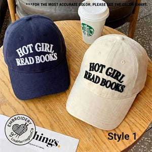 May include: Two baseball caps, one navy blue and one cream, are displayed on a wooden surface. Both caps feature the embroidered text "HOT GIRL READ BOOKS" in white and black, respectively. A coffee cup with a green logo is visible.