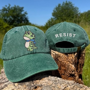 May include: Two green baseball caps. One cap features an embroidered frog with a blue scarf. The other cap has the word "RESIST" embroidered on the back. Both caps are on a wooden surface, with a blurred green background.