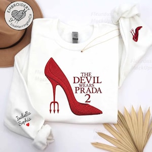May include: White sweatshirt with a red high-heeled shoe design and the text "THE DEVIL WEARS PRADA 2." The sleeve is embroidered with the name "Isabella Sophia." A small trident is embroidered on the other sleeve.