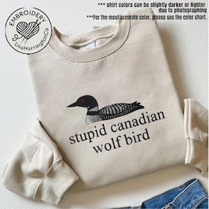 Pode incluir: Um moletom bege com um desenho de mergulhão bordado em preto e branco e o texto "stupid canadian wolf bird". O moletom também apresenta o logotipo "Embroidery LisaHarringtonCa".