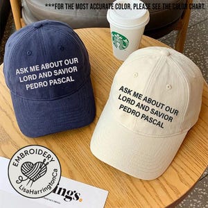 May include: Two baseball caps, one navy blue and one off-white, are displayed. Both caps feature the embroidered text: "ASK ME ABOUT OUR LORD AND SAVIOR PEDRO PASCAL." A coffee cup is visible in the background. The caps are on a wooden surface.