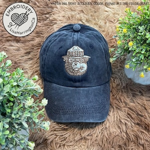 May include: A black denim baseball cap with a gold embroidered image of a bear wearing a ranger hat. The bear is wearing a hat with the word "RESIST" on it.