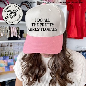 May include: A white and pink baseball cap with the text "I DO ALL THE PRETTY GIRLS' FLORALS". The cap has a curved brim and a structured crown. The cap also has the logo of "Embroidery LisaHarringtonCa" on the side.