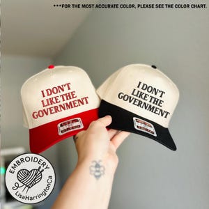 May include: Two trucker hats with the text "I DON'T LIKE THE GOVERNMENT". One hat has a red brim and the other has a black brim. Both hats have a cream-colored crown and a small tag.