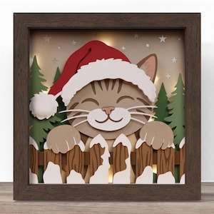 May include: A layered paper shadow box art piece featuring a brown tabby cat wearing a red and white Santa hat. The cat rests its paws on a brown wooden fence covered in white snow. Green pine trees and small white stars are in the background.