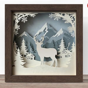 Könnte beinhalten: Gerahmtes Papierkunstwerk, das eine Winterlandschaft darstellt. Das Design zeigt einen weißen Hirsch, Häuser, Bäume und Berge, vor einem blau-grauen Hintergrund. Der Rahmen hat eine dunkelbraune Farbe.