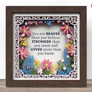 May include: A framed paper art piece with a brown wooden frame. The artwork features a layered floral design in pink, blue, yellow, and white, surrounding the text "You are BRAVER than you believe, STRONGER than you seem and LOVED more than you know."