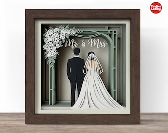 May include: A framed paper art piece featuring a bride and groom under an archway with the words "Mr & Mrs." The artwork is layered with white roses and greenery, set in a brown wooden frame. The bride wears a white wedding dress and veil.