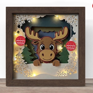 Op de afbeelding: Een ingelijst, gelaagd papierkunstwerk met een cartoon eland in een winters tafereel. De eland heeft grote ogen en een gewei. De scène omvat kerstbomen, sneeuwvlokken en rode ballen. Warmwitte lichten verlichten het kunstwerk.