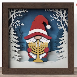 May include: A framed paper art piece featuring a gnome holding a menorah. The gnome wears a red hat and holds a gold menorah with lit candles. The background depicts a winter scene with white trees and snow.