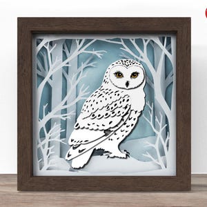 May include: A framed, layered paper art piece featuring a snowy owl. The owl is white with black spots and yellow eyes, set against a backdrop of a winter forest with white trees and a blue sky. The frame is a dark brown.