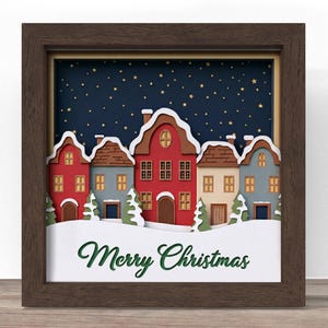 May include: A framed Christmas artwork depicting a village scene with red, blue, and beige houses under a starry night sky. The houses have snow-covered roofs against a dark blue backdrop. The words "Merry Christmas" are written in green.