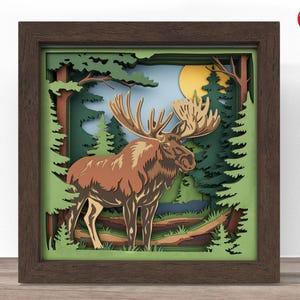 May include: A framed paper art piece featuring a brown moose with large antlers in a forest scene. The artwork uses layered paper to create depth, with a yellow sun and green trees. The frame is dark brown, and the artwork is labeled "Crafter Lobby".