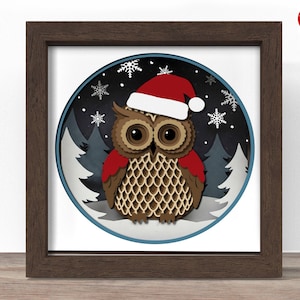 May include: A framed paper art piece depicting a brown owl in a red Santa hat, set against a dark blue, snowy backdrop with pine trees and snowflakes. The frame is dark brown.