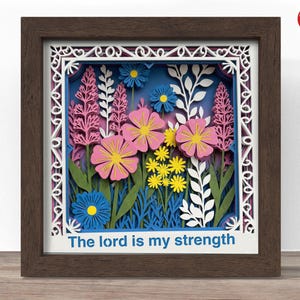 Könnte beinhalten: Ein gerahmtes Papierkunstwerk mit einem mehrschichtigen Blumendesign. Rosa, gelbe und blaue Blumen vor blauem Hintergrund. Der Text "The lord is my strength" steht unten. Der Rahmen ist braun.