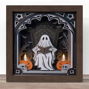 May include: A layered paper shadow box in a dark brown frame featuring a white ghost sitting on a throne reading a book. The scene includes two orange pumpkins, two white skulls, and two small lit candles against a black background with bats.