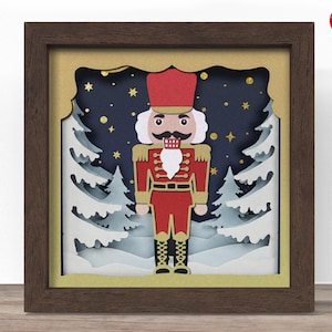 May include: A framed, layered paper art piece featuring a red and gold nutcracker soldier. The artwork includes white trees, a starry night sky, and gold accents. The frame is a dark brown color, and the artwork is set against a gold background.