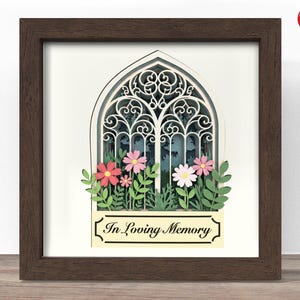 Op de afbeelding: Een ingelijst papieren kunstwerk met een kerkraamontwerp. Het witte raam heeft gedetailleerde uitsparingen tegen een blauwe achtergrond. Papieren bloemen in roze en wit en de tekst "In Loving Memory" staan onderaan het kunstwerk.
